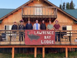 Seven men stand on the wooden porch of Lawrence Bay Lodge with a large sign in front, facing the camera.