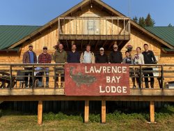Ten people stand on a wooden deck in front of a sign reading "Lawrence Bay Lodge" at a rustic lodge building.