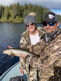 Two people in camouflage jackets pose on a boat, holding a freshly caught fish, with a forested lake in the background.