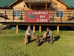 Two people kneel on grass holding moose antlers in front of a wooden lodge with a "Lawrence Bay Lodge" sign.