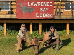 Two people kneel on grass holding a large moose antler rack in front of a Lawrence Bay Lodge sign.