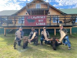 Four people kneel with large moose antlers in front of Lawrence Bay Lodge, a wooden building with a red sign.