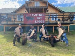 Four people kneel in front of two large moose antlers outside Lawrence Bay Lodge, a wooden lodge building.