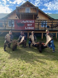 Four people kneel in front of a lodge holding two large moose antlers, with a sign reading "Lawrence Bay Lodge" behind them.