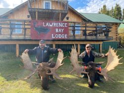 Two people kneel with large moose antlers in front of Lawrence Bay Lodge, a wooden building with a red sign.