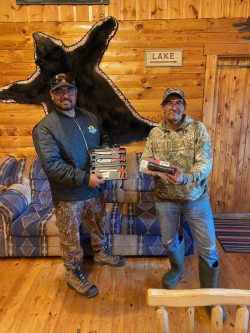 Two men stand indoors holding boxes; a bear rug and a "LAKE" sign hang on the wood-paneled wall behind them.