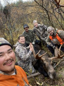 Five hunters in camouflage pose with a harvested moose in a wooded area during autumn.