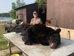 A man in camouflage clothing poses behind a large, black bear lying on a table outdoors near a wooden building.