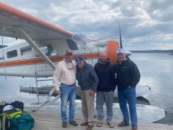 Four men stand on a dock in front of a floatplane by a lake, with cloudy skies in the background.
