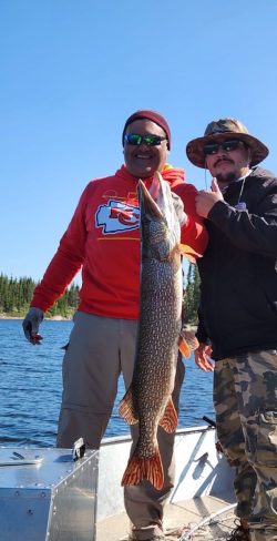 Two men on a boat hold up a large fish, with a forested shoreline and blue water in the background.
