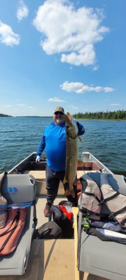 Person standing on a boat holding a large fish upright with one hand, with lake and trees in the background.