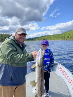 Two people on a boat show off a large fish they caught on a lake surrounded by trees under a partly cloudy sky.