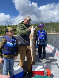 Two children and an adult on a boat display a large fish they caught, with a lake and trees in the background.