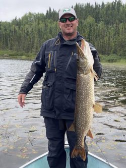 Person standing in a boat holding a large fish in front of a lake with trees in the background.