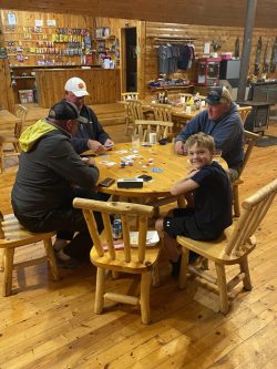 Four people sit around a wooden table playing cards in a rustic room; one child looks at the camera and smiles.