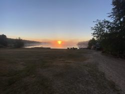 A sunrise over a calm lake with boats on the shore and a grassy, sandy area in the foreground.