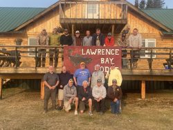 A group of people pose in front of a wooden lodge with a sign that reads “Lawrence Bay Lodge.”.