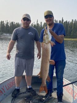 Two men on a boat hold a large fish with trees and water in the background.