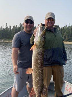 Two men on a boat hold up a large fish they caught, with a forested shoreline visible in the background.