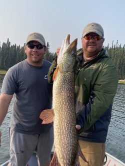 Two men on a boat hold a large fish in front of them with a lake and trees in the background.