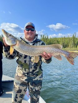 Man in camouflage clothing holds a large northern pike fish on a boat, with water and trees in the background.