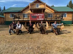 Four people kneel outside Lawrence Bay Lodge holding large moose antlers in front of a wooden building.