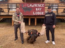 Two people stand outside Lawrence Bay Lodge holding large moose antlers attached to a moose head on the ground.