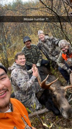 Five hunters pose with a moose they have hunted in a forested area, smiling at the camera.