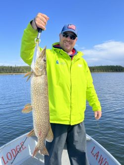 Man in a neon yellow jacket holds up a large fish on a boat in a lake, with trees and sky in the background.