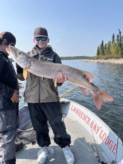 A person in a jacket and cap holds a large fish on a boat with "Lawrence Bay Lodge" written on the side.
