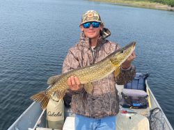 Person in camouflage jacket and hat holding a large fish on a boat in a calm lake.
