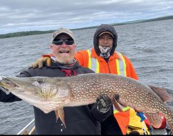 Two men on a boat hold a large northern pike fish, with a choppy lake and cloudy sky in the background.