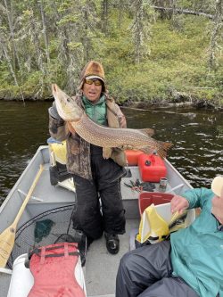 Person standing in a boat holds a large fish, while another person sits nearby; forested riverbank in background.