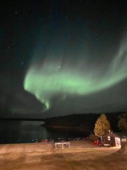 Green aurora borealis arcs across the night sky above a lakeshore with a small building and boats in the foreground.