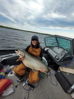 Person sitting in a boat holding a large fish, with fishing gear and water visible in the background under a cloudy sky.
