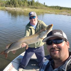 Two men on a boat; one holds a large fish while the other takes a selfie on a lake with trees in the background.