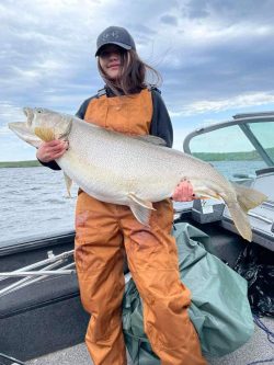 Person wearing orange overalls holds a large fish on a boat with water and a cloudy sky in the background.
