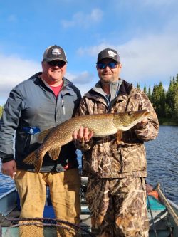 Two men on a boat hold a large Northern pike fish with a lake and trees in the background.