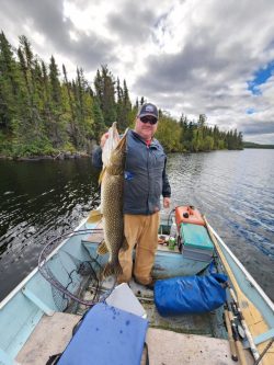 Man standing in a small boat on a lake holding a large fish with trees and cloudy sky in the background.