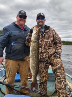 Two men on a boat pose with a large fish they caught; one man holds the fish vertically by its mouth.