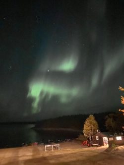 Green aurora borealis lights swirl in the night sky over a lakeside cabin and trees.