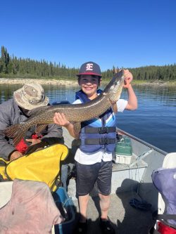 Smiling boy in a life jacket holds a large fish on a boat, with another person sitting and trees in the background.