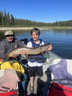 A boy on a boat holds a large fish, smiling, with an adult beside him; trees and water in the background.