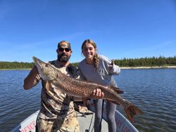 Two people on a boat hold a large fish together on a lake with trees and blue sky in the background.