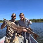 Two people on a boat hold a large fish together on a lake with trees and blue sky in the background.