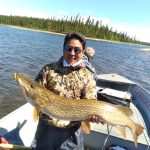 Person standing on a boat holding a large fish with a lake and forested shoreline in the background.