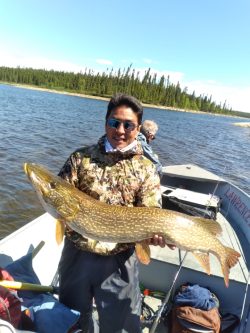 Person standing on a boat holding a large fish with a lake and forested shoreline in the background.
