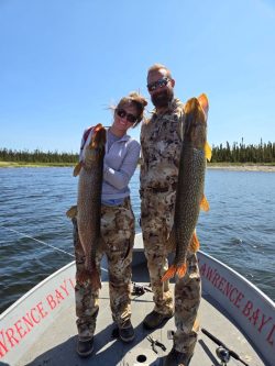 Two people in camouflage clothing stand on a boat holding large fish, with water and trees in the background.