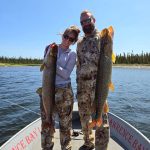 Two people in camouflage clothing stand on a boat holding large fish, with water and trees in the background.