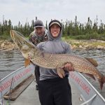 Two people on a boat; the person in front holds a large fish while the other stands behind, smiling.